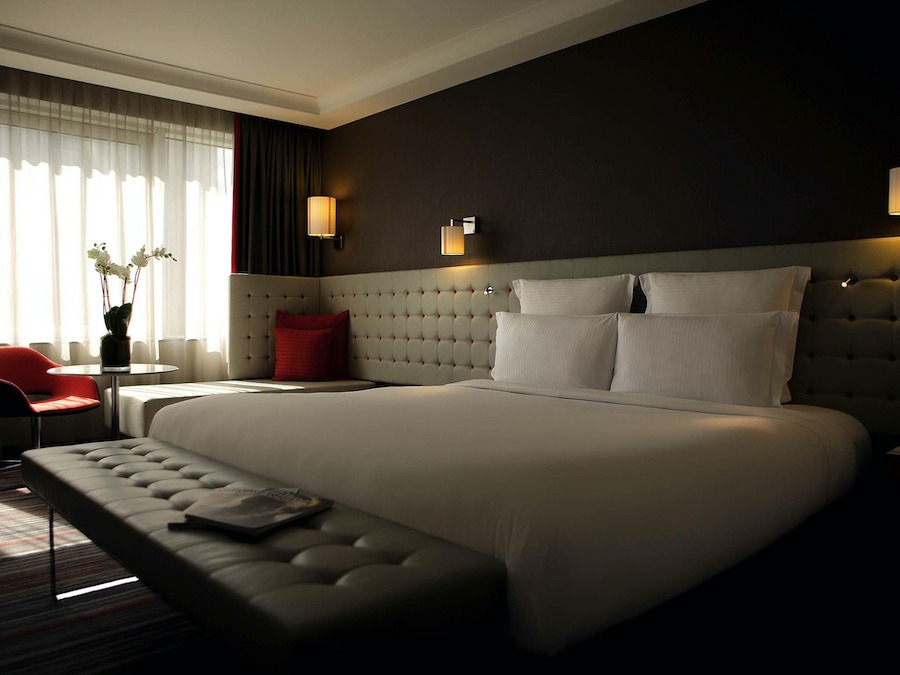 This is an image of a cool hotel room with a neatly made double bed and sleek furniture and low lighting and dark walls.