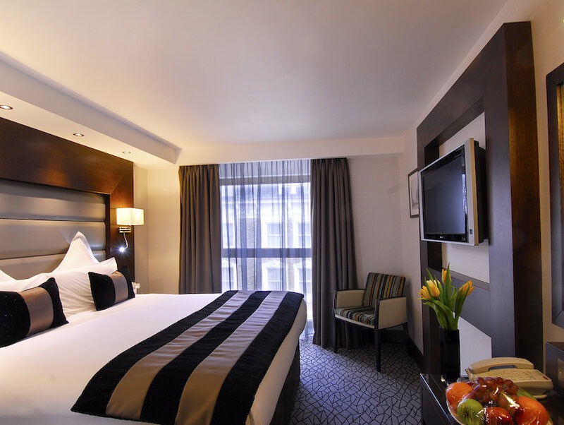 This is an image of a cool hotel with a big double bed that is neatly made, windows with dark grey curtains, a grey carpet, wooden furnishings, soft lighting, a grey armchair in the corner of the room and a television that is hung on the wall opposite the bed. There are also flowers underneath the television screen and fruit on the table opposite the bed.