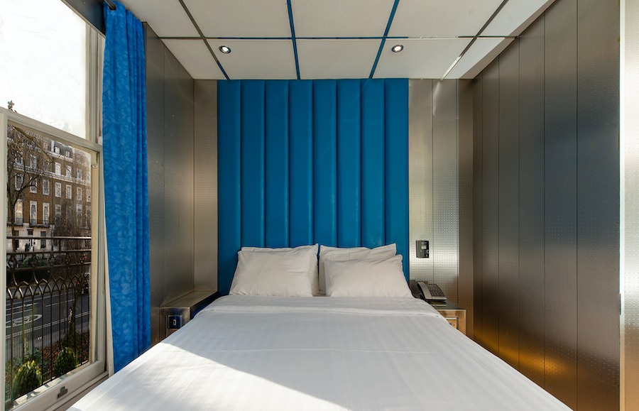 This is an image of a small hotel bedroom with a neat white double bed, a window, bright blue furniture detailing and minimalistic furnishings. 