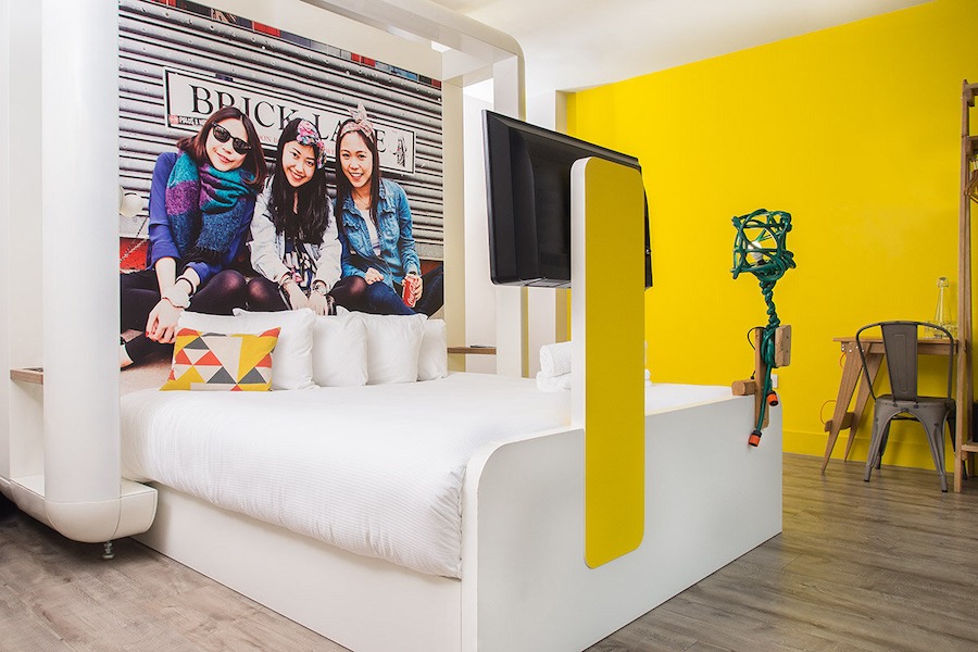 This is a photo of a bright hotel bedroom. It has a bright yellow wall and wooden floors and the bed is neat and has white sheets and bedding. There is a cool poster behind the head of the bed of three people smiling with the BRICK LANE sign behind them. The room has a trendy, cool vibe with a funky aesthetic. 