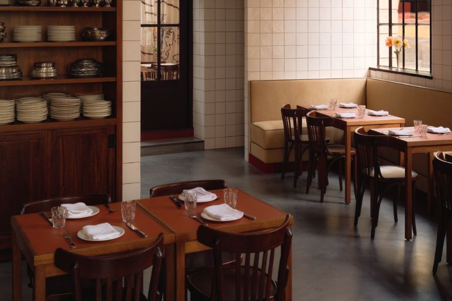 The interior of a relaxed bistro-style restaurant with warm wooden tables set with white napkins and glassware, bentwood chairs, cream banquette seating, white subway tiles, and open shelving displaying stacked crockery.