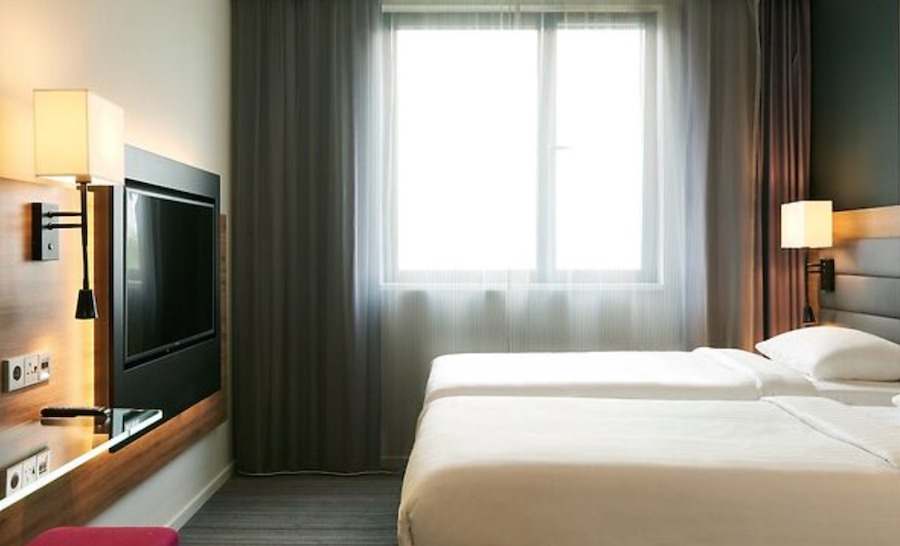 This is an image of a hotel bedroom with two single beds neatly made with white sheets and minimalistic furnishings. There is a flat screen tv on the wall opposite the beds. 