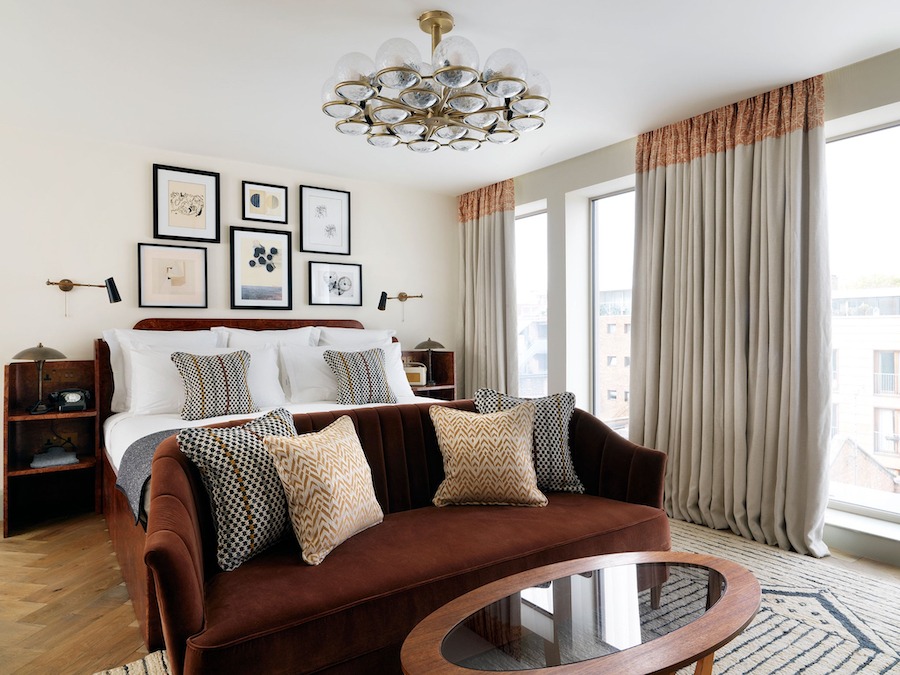 This is an image of a big hotel room with a double bed, sofa, table and small framed artworks that are hung up on the wall behind the bed. The room is toned in muted colours, such as grey, taupe and beige. It is classy and elegant and minimalistic yet warm.