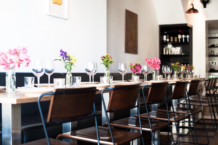 This is an image of a restaurant with wooden tables and chairs all in one line. There are colourful flowers on the table and the aesthetic is clean and minimalistic.