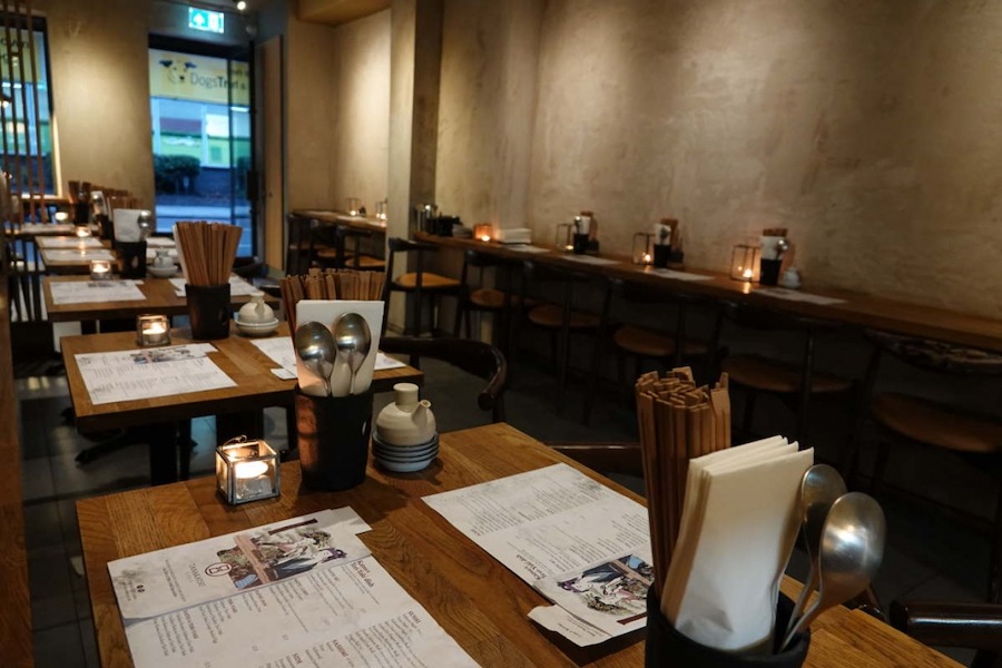 This is an image of a dimly lit restaurant. There are square wooden tables in the room with candles and cutlery on the tables. The walls are bare and it is minimalistic.