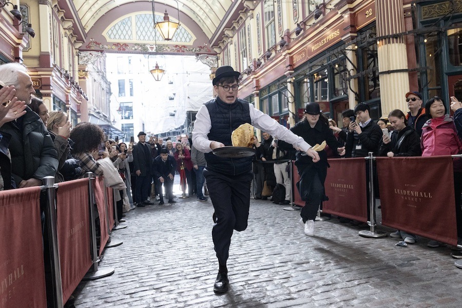 This is an image of a man flipping pancakes running down a street with spectators watching.