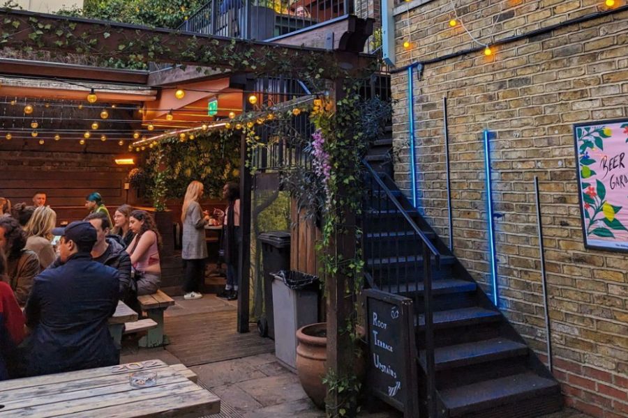 This is an image of a outdoor beer garden with people scattered across wooden tables talking and drinking. There are fairy lights adorning the roof.