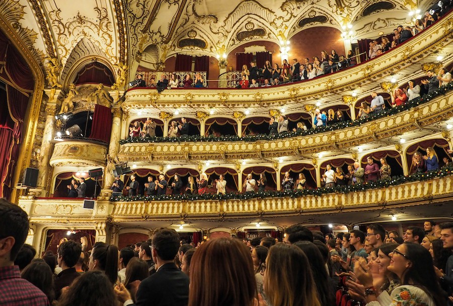 This is an image of a crowded and full theatre with lots of balconies and lights. The crowd appears to be clapping.