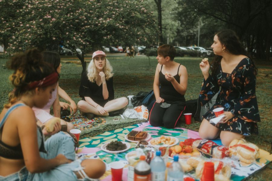 This is a picture of a group of friends having a picnic together in the park and laughing. They have a bunch of food in front of them.