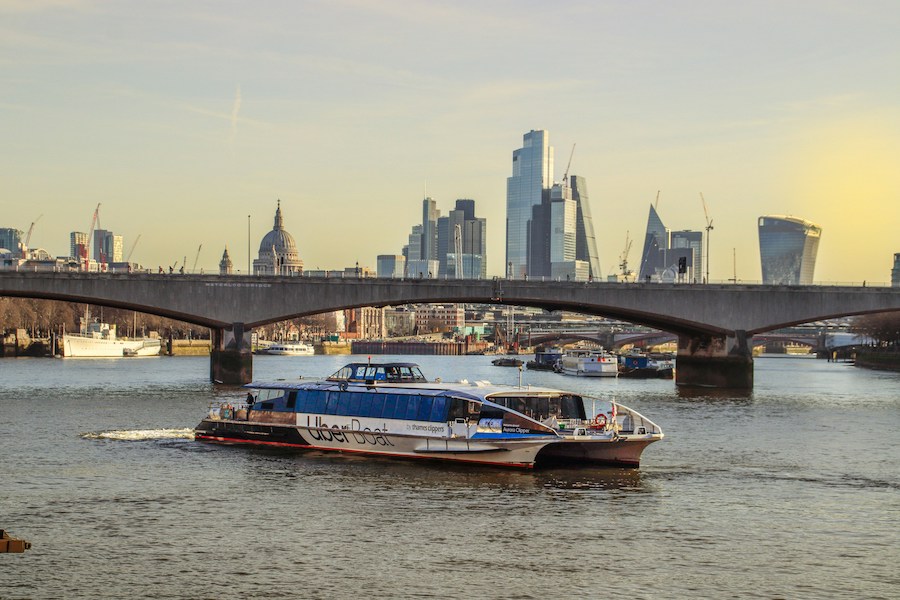 This is an image of an Uber boat gliding underneath a bridge. The city of London can be seen behind it. A hop-on-hop-off bus is one of London's most overrated attractions. Try Uber Boat instead.