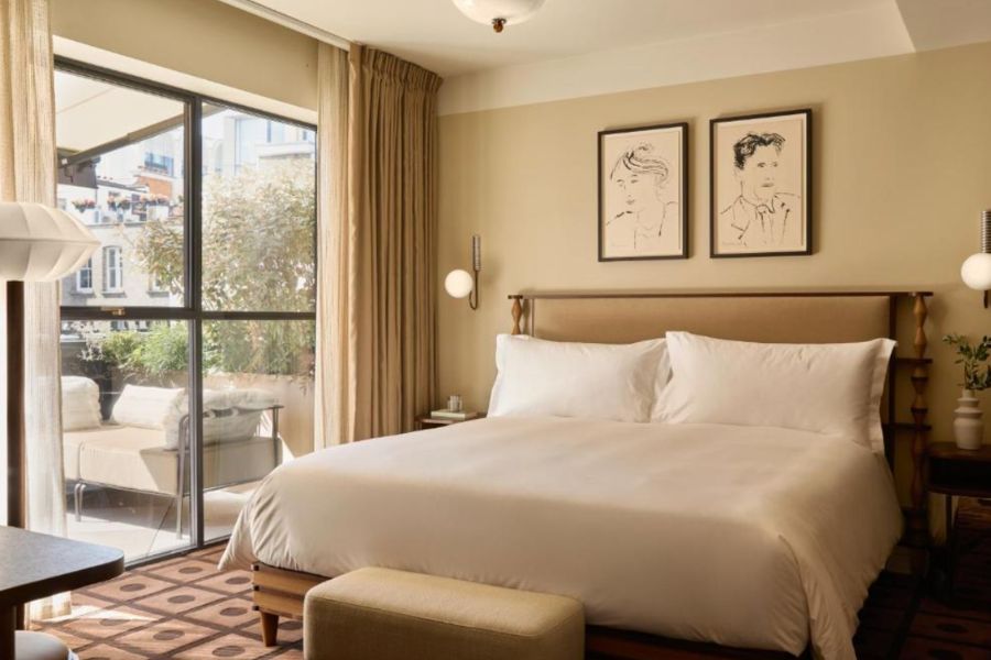 A bright, airy hotel room with a king-size bed dressed in white linen, neutral sand-toned walls, two framed portrait sketches above the headboard, globe wall sconces, a geometric-patterned rug, and a large glass door opening onto a private balcony with a seating area.