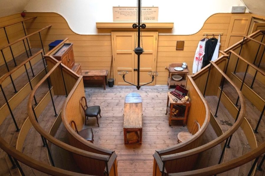 An overhead view of a historic Victorian operating theatre, featuring a curved wooden viewing gallery, a central wooden operating table, surgical instruments on a side trolley, a blood-stained surgeon's apron hanging on the wall, and a skylight overhead.