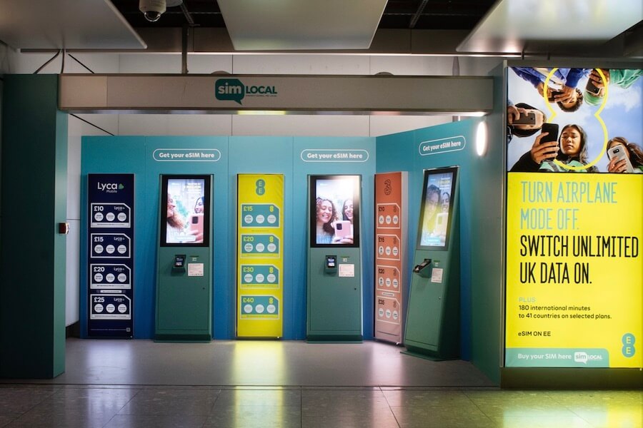 Here's alt text for the image: "A row of three self-service eSIM kiosks at a SimLocal stand in what appears to be an airport or transit hub. The kiosks represent three UK mobile networks — Lyca Mobile (teal), EE (yellow), and Three (coral/orange) — each displaying pricing plans in pounds and touchscreens showing customers using phones. A yellow EE advertisement to the right reads 'Turn Airplane Mode Off. Switch Unlimited UK Data On.' The booth is set against a teal backdrop with 'Get your eSIM here' signage above each kiosk.