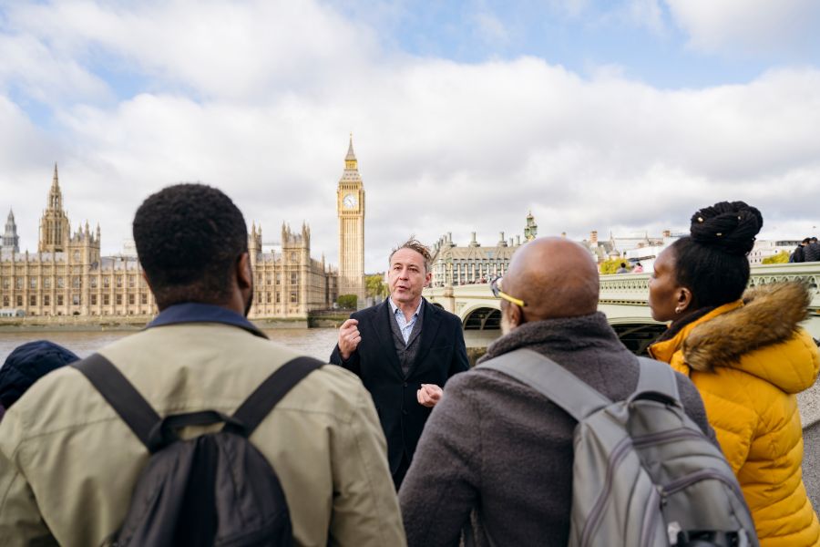 This is an image of a man taking a tour group around London. They are currently across the Thames with Big Ben in the distance behind them.