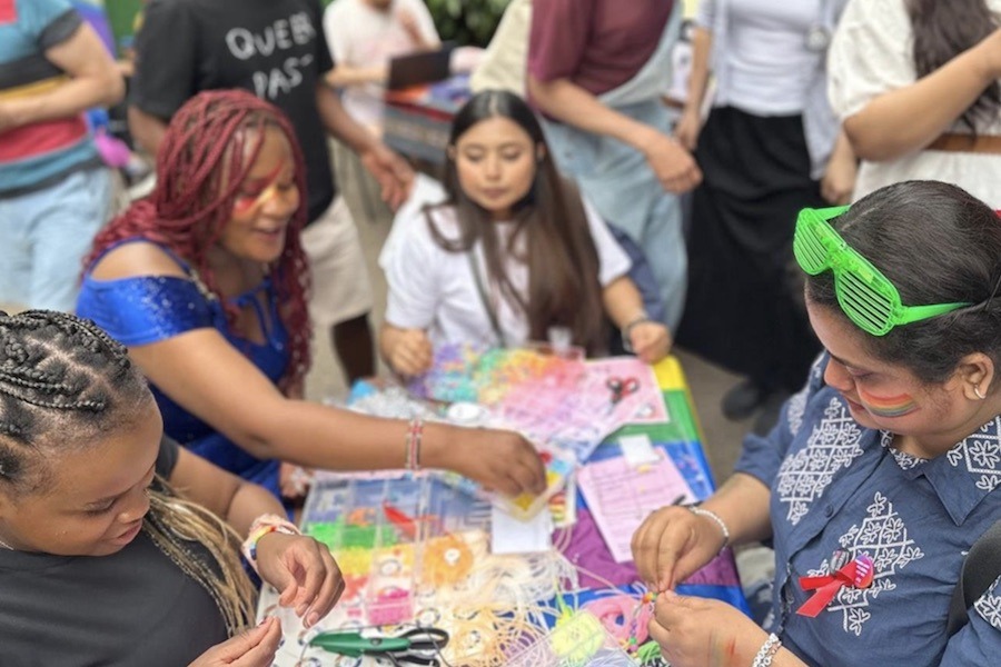 People gathered around a colorful craft table at an outdoor festival, making friendship bracelets and other crafts. Several attendees have rainbow face paint, and one person wears novelty green sunglasses on their head. A t-shirt in the background reads "Queer" and "Past."