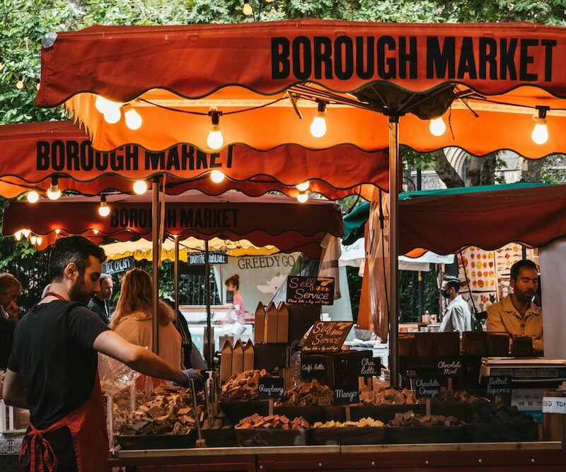 This is a picture of a vendor at Borough Market. His small has a bright orange Borough Market branded umbrella on the top of it.