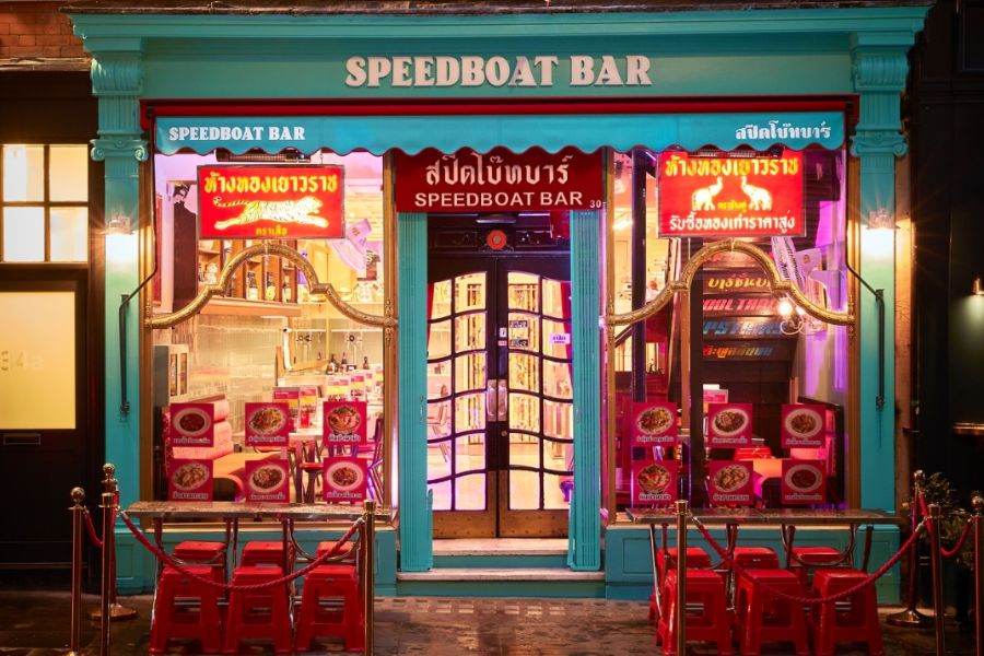 The vibrant neon-lit exterior of Speedboat Bar at night, with a teal-painted facade, Thai-language signage, glowing red neon signs, red plastic stools on the pavement outside, and red rope barriers.