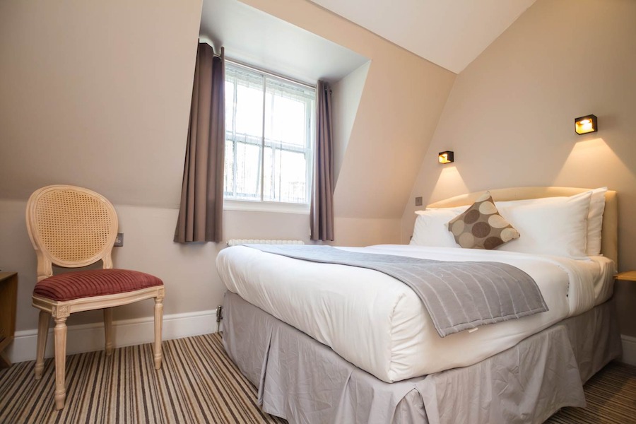 This is an image of a clean hotel bedroom toned in muted colours of beige white grey and brown. It is minimalistic and elegant with a bed, window and chair.