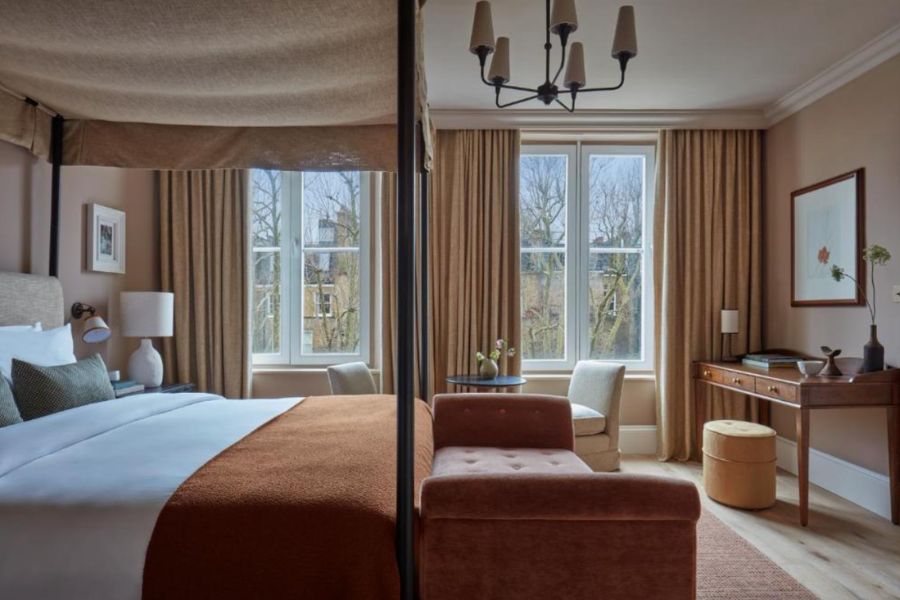 A warmly decorated hotel room with a four-poster bed, burnt orange throw, bay windows overlooking bare winter trees, a chandelier, a velvet chaise longue, a wooden writing desk with a mirror, and soft terracotta and neutral tones throughout.