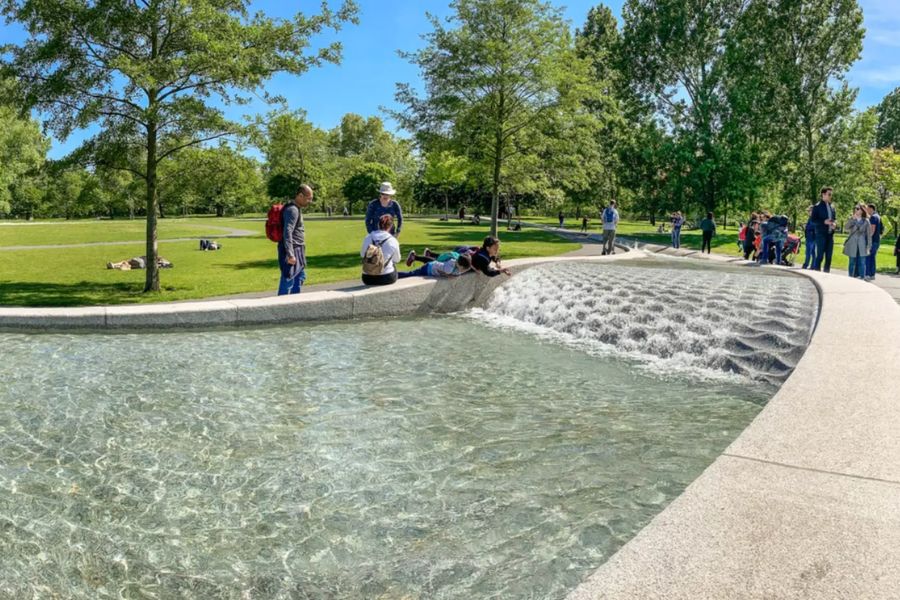 This is an image of a water fountain in the middle of Hyde Park. There are kids on the side leaning into the water, while their parents watch from the sidelines.