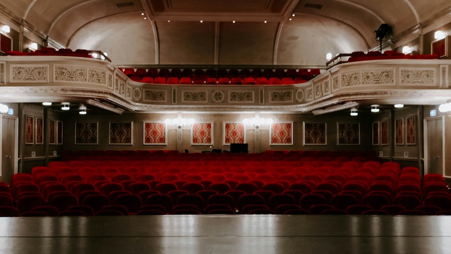 This is a photo of the inside of a theatre lined with rows of red chairs. There is a floor level and a dress circle above.