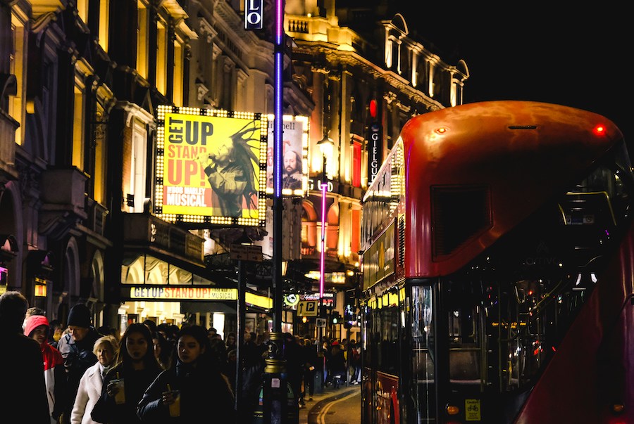 This is a picture of a busy West End London street at night. There are lots of people around, as well as a big, red, double-decker bus riding alongside them. One of the things to know before booking London theatre tickets is that you should watch out for people selling scam tickets on the street in Picadilly Circus or other tourist areas.