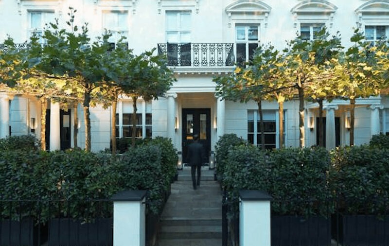 This is an image of a stylish front of a white hotel with trees and hedges in the courtyard.