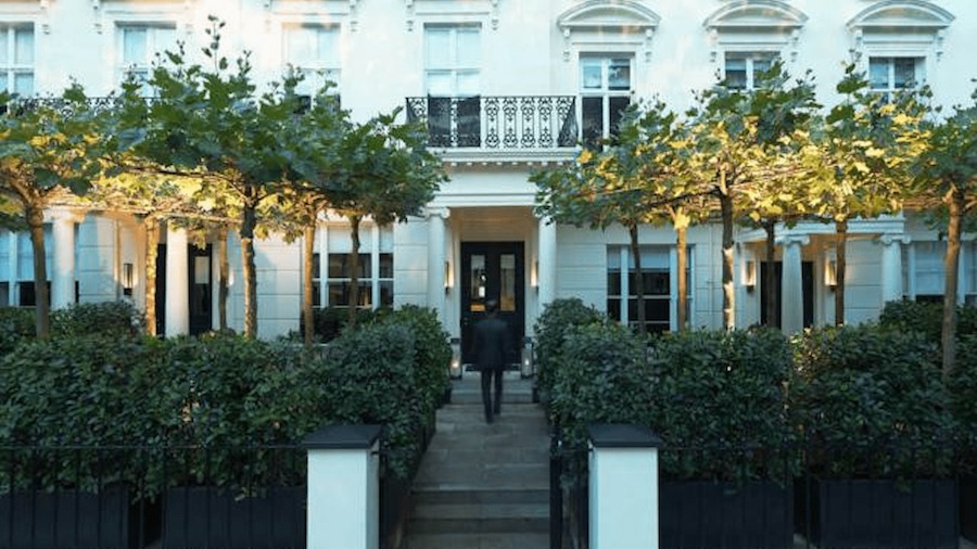 This is an image of a stylish front of a white hotel with trees and hedges in the courtyard. 