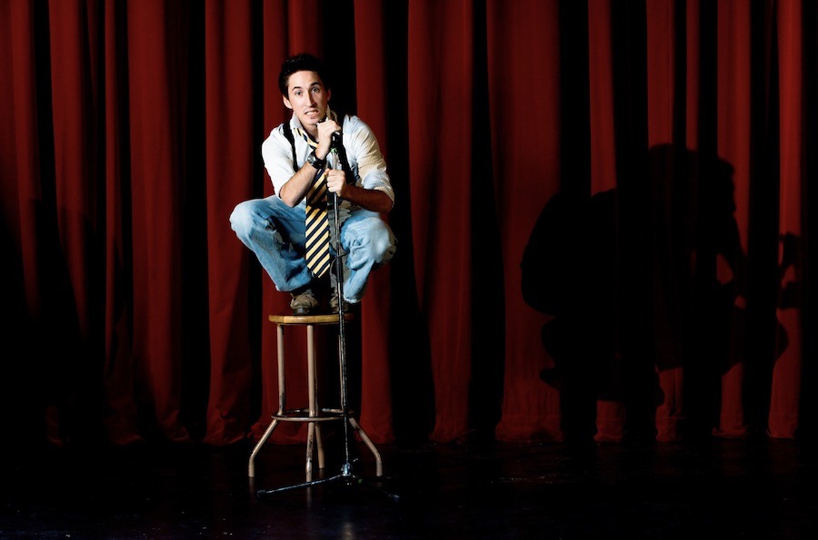 This is an image of a comedian sitting on a stool on stage with a mic. There is a red curtain closed behind him.