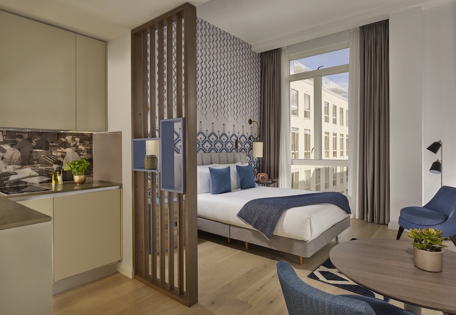 This is an image of a big hotel bedroom that is modern, sleek and luxurious with muted blue details to the furnishings. It is elegant and classic in style.