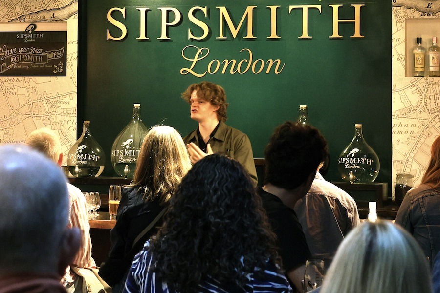 A guide speaks to a seated group during a Sipsmith gin tasting, with the Sipsmith London sign and large glass demijohns displayed on the wall behind him.