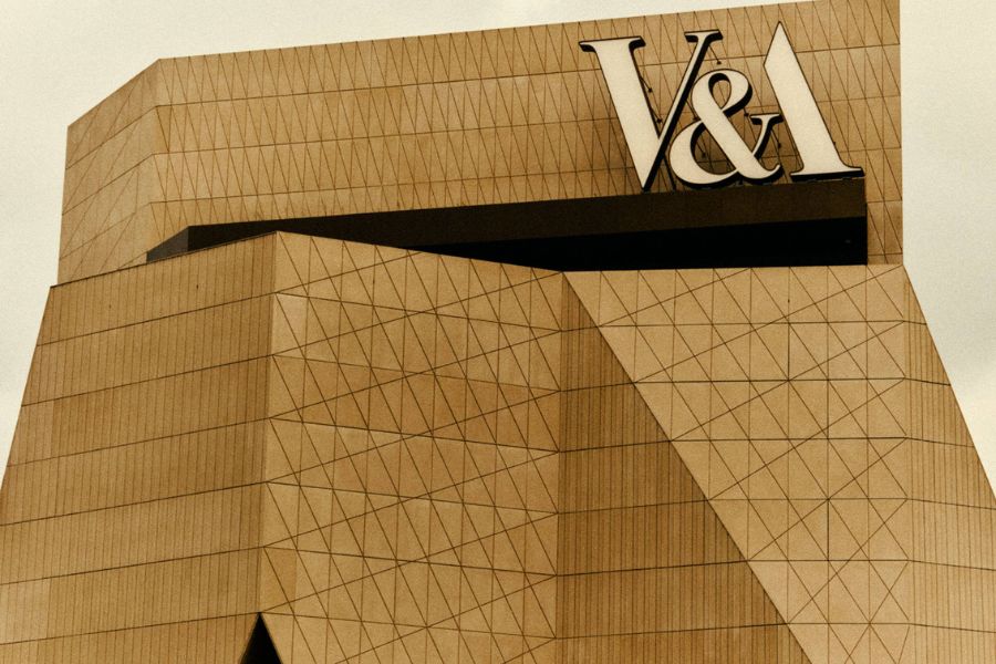 A close-up architectural shot of the V&A East Museum building, showing its dramatic angular golden facade clad in a geometric triangular pattern, with the bold V&A logo displayed at the top.