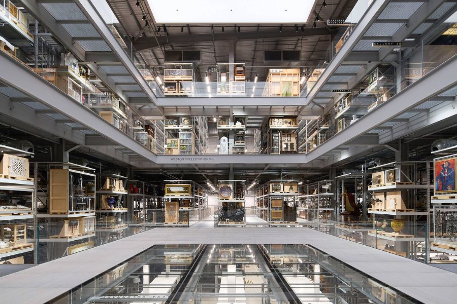 he vast interior of the V&A East Storehouse (Weston Collections Hall), showing multiple open floors of glass-fronted storage shelving densely packed with artworks, artefacts, and objects from the museum's collection, with a reflective glass floor below.