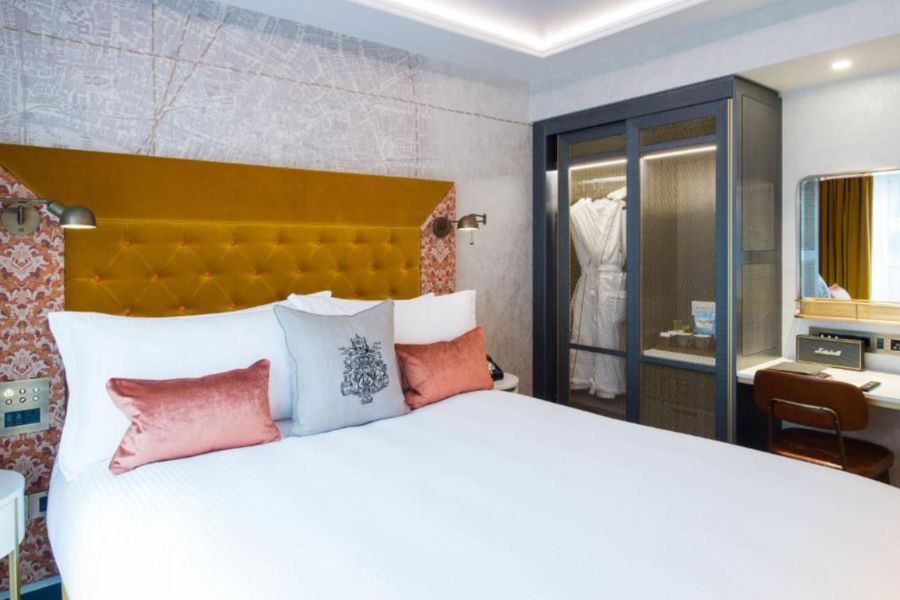 A hotel guest room at Vintry & Mercer featuring a king-size bed with a mustard yellow tufted velvet headboard, pink and grey accent cushions, patterned wallpaper, a glass-fronted wardrobe with robes visible, and a Marshall speaker on the desk.