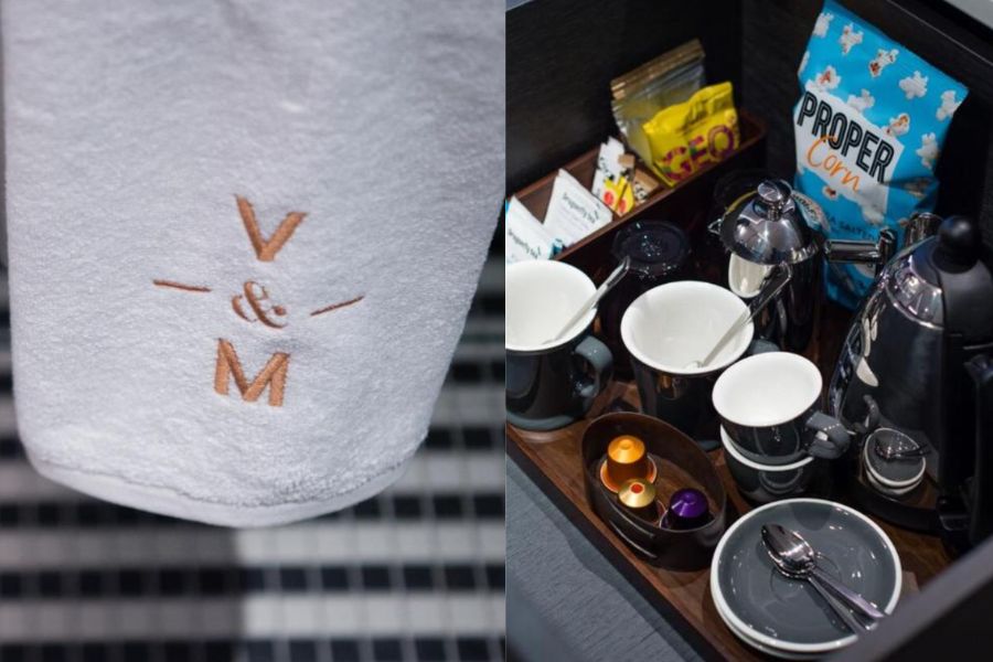 Close-up of a white towel embroidered with the V&M logo, alongside an overhead view of the in-room hospitality tray featuring a Nespresso machine, mugs, tea, and snacks including Proper Corn popcorn.