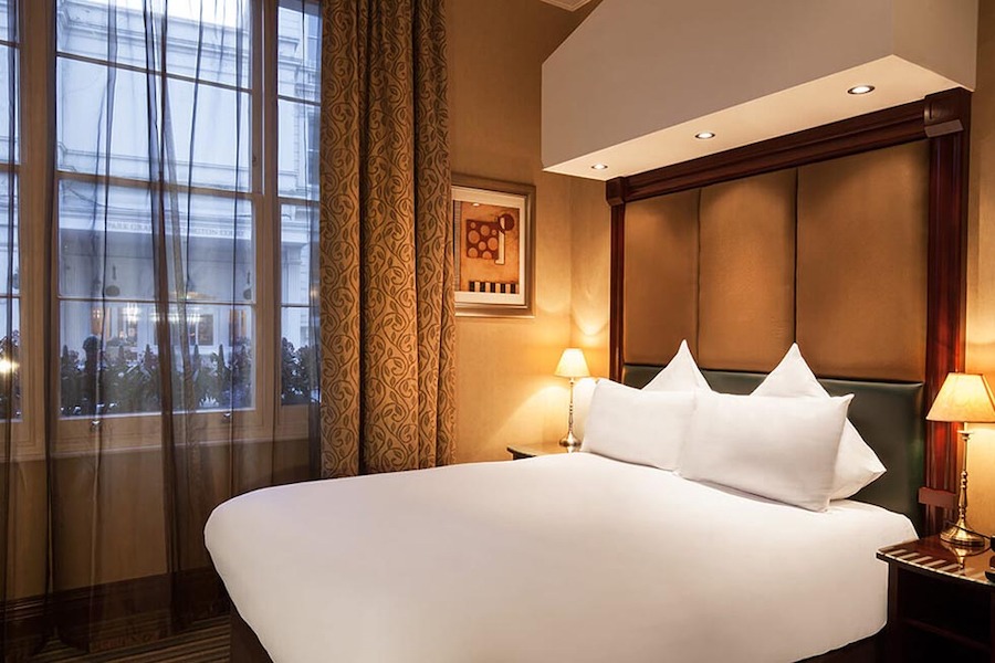 This is an image of a softly lit hotel bedroom. The bed is white and impeccably made up. The room is toned in muted colours of brown, beige, white and grey. 