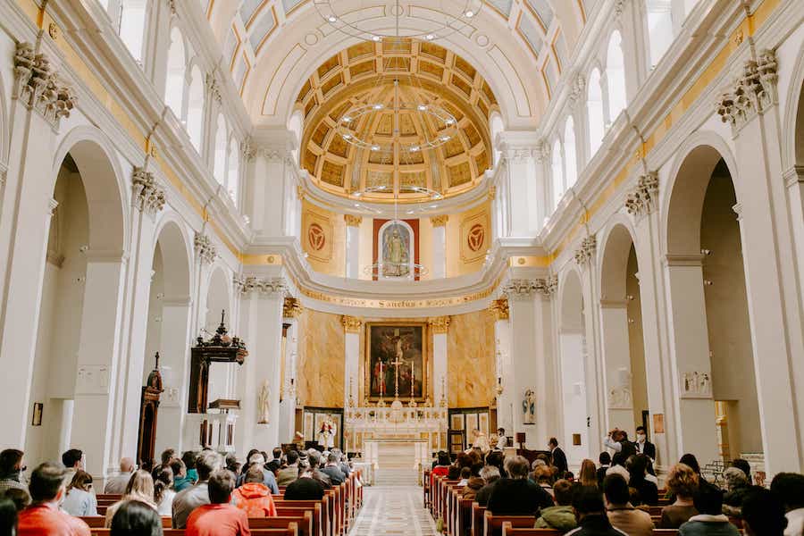 This is an image of a beautiful and well lit catholic church in London. There are high archways either side of the room and a large group of people is sat in the aisles ready to listen to the service. The altar is toned in rich gold colours that gives the space a regal aesthetic.