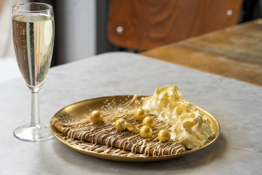 This is an image of a crepe on a gold plate. There is a glass of champagne next to the plate on a grey table. The pancake is drizzled with cream and is topped with gold circular chocolate balls.