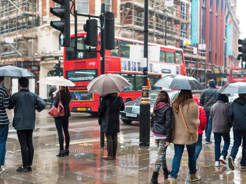 Why people might not enjoy their trip to London: They didn't plan for the weather. This image is of a bunch of people holding their umbrellas in the rain. There is a double-decker bus in the background.
