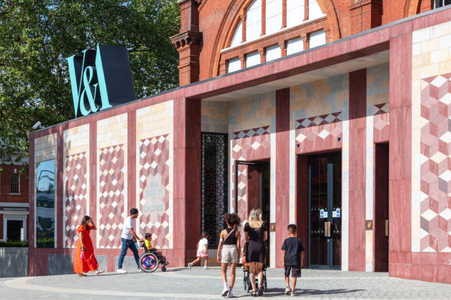 The entrance to the Young V&A (V&A Museum of Childhood) in Bethnal Green, featuring a colourful refurbished Victorian red-brick facade with decorative geometric tilework in shades of red, pink, and white, a teal V&A logo, and visitors including families with young children approaching the entrance on a sunny day.